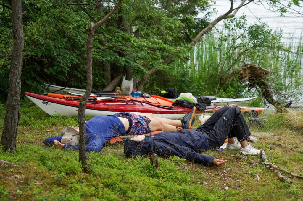 Three kayaks by the shore on a mossy island.  
The paddlers resting in the grass beside them.